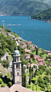 Vertical view of Santa Maria del Sasso in Morcote, Switzerland, highlighting its historic bell tower above hillside homes with the deep blue waters of Lake Lugano behind.