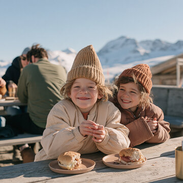 Two Cute Smiling Kids in Knit Beanies Sitting at Outdoor Table with Burgers in Mountains