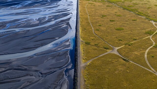 Aerial view of the Markarfljot glacial river featuring a stark contrast between dark braided channels and green plains separated by a straight embankment, Rangarthing eystra, Iceland.