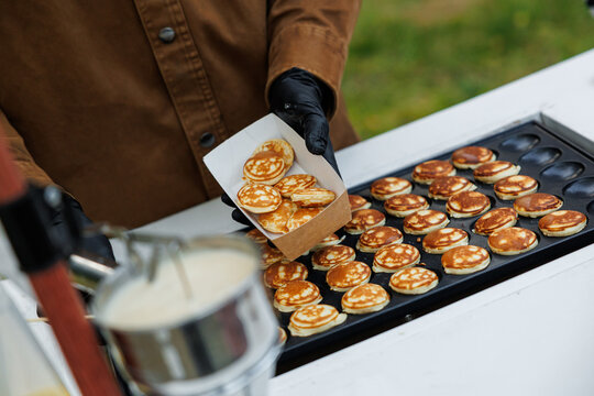 Gloved vendor places freshly cooked mini pancakes into a paper tray beside a hot griddle and bowl of batter at an outdoor food stand