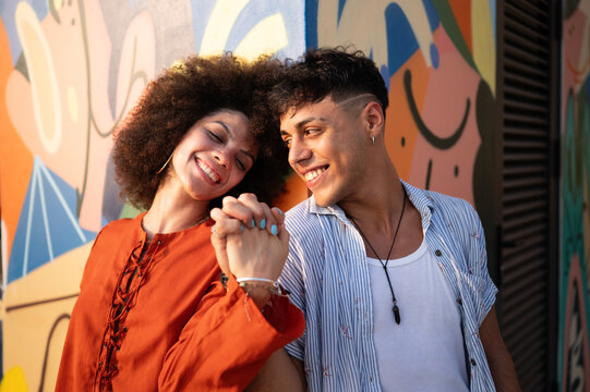 Joyful man and woman looking at each other with love while holding hands against a bright wall