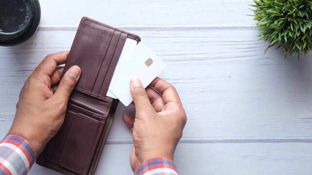 Hands inserting credit card into brown leather wallet on wooden surface