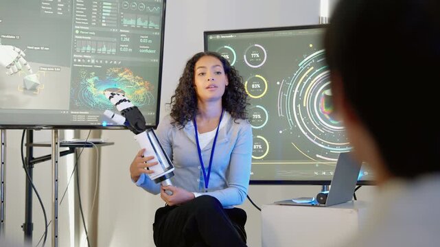 Female engineer presenting a robotic arm prototype to colleagues in a technology research environment, with digital displays showing robotics data and performance analysis.
