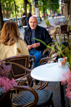 A bald middle-aged man and a woman talking on the patio of a caf&eacute; on the street