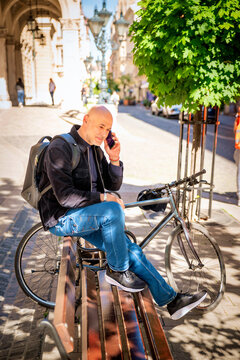 A middle-aged man sitting on a bench in a European city, next to his bicycle, talking on the phone