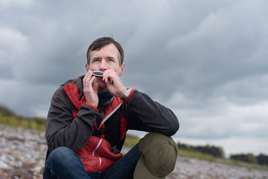 A thoughtful man plays a harmonica while seated on rocky beach, surrounded by moody clouds and coastal landscape. The man has a new hobby, he is learning to play the harmonica