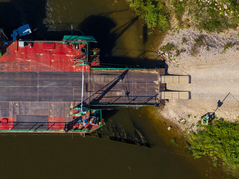 Aerial view of the ferry in Vilkija docked at the riverbank with a metal ramp, red and green deck, and gravel shore in Vilkija, Kaunas County, Lithuania.