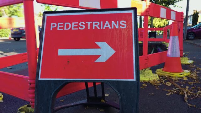 Pedestrian direction sign beside roadworks barriers on sidewalk. White arrow on red temporary safety sign near traffic cone. Street construction area with pedestrian route marker and red plastic