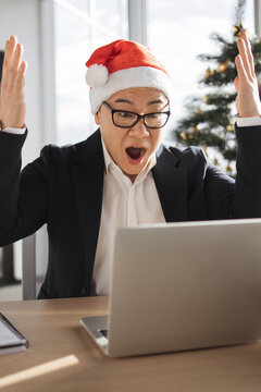 An astonished Asian man wearing a Santa hat and glasses reacts with shock while looking at his laptop screen during the holiday season