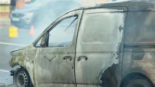 Burned van smoking on urban roadway after vehicle fire. Fire damaged vehicle with charred bodywork, shattered windows and scorched panels after blaze. Smoking car wreck on wet street with traffic