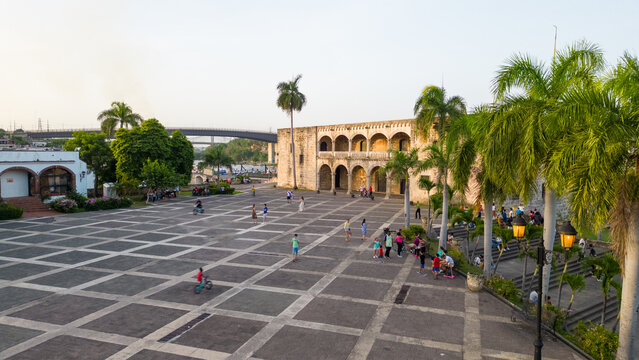 Aerial view of the historic Alcazar de Colon in the Zona Colonial, featuring a large stone-paved plaza, palm trees, and people walking, Santo Domingo, Distrito Nacional, Dominican Republic.