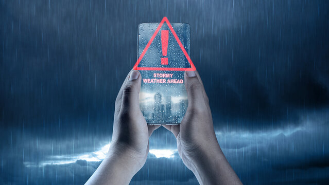 Hands holding a smartphone displaying a red warning alert for stormy weather against a rainy background. Concept of Weather