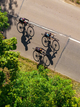 Aerial view of three cyclists riding on an asphalt road with long shadows cast on the pavement, bordered by green trees in Vilnius, Vilnius County, Lithuania.