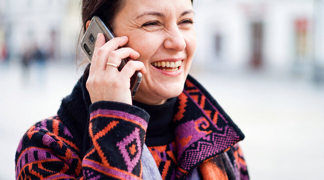 A genuinely smiling adult woman in bright patterned clothes enjoys talking on a mobile phone on the street. Talking on the phone