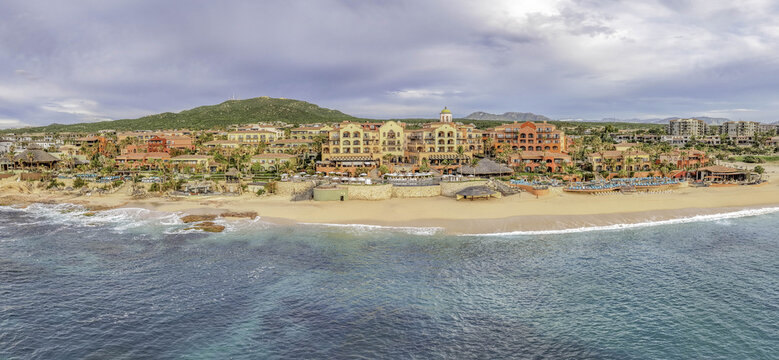 Aerial view of Hacienda Del Mar resort complex with terracotta-colored buildings, palm trees, and swimming pools along the sandy beach and turquoise ocean in Cabo, Mexico.