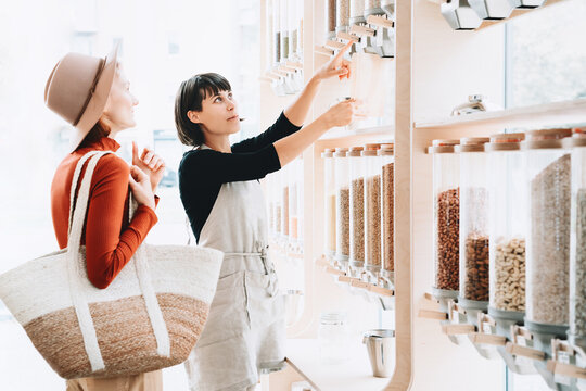A female shopkeeper in an apron assists a young woman in using gravity food dispensers in a modern shop. Conscious consumer behavior and package free shopping concept.