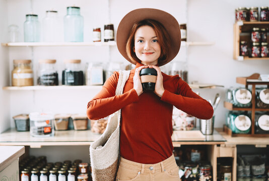 A stylish caucasian female customer wearing a hat holds a reusable coffee cup inside a bulk grocery shop. Mindful lifestyle and conscious environmental consumerism concept.