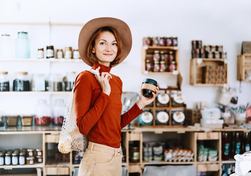 A stylish caucasian female customer wearing a hat holds a reusable coffee cup inside a bulk grocery shop. Mindful lifestyle and conscious environmental consumerism concept.