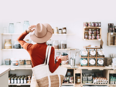 Shopper stands with her back to the camera, looking at organic goods on wooden shelves inside a bright modern store interior. Package free retail environment and conscious consumption concept.