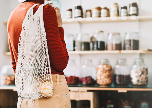 Shopper stands with her back to the camera, looking at organic goods on wooden shelves inside a bright modern store interior. Package free retail environment and conscious consumption concept.