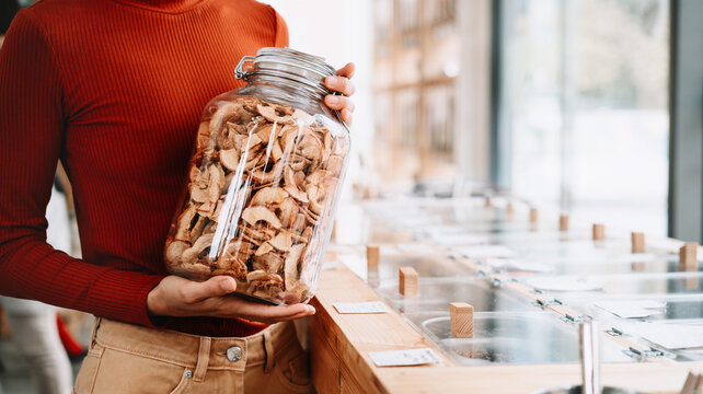 A happy caucasian female customer in a terracotta sweater holds a large glass jar of dried apples inside a bulk shop. Sustainable consumer choices and low waste lifestyle concept.