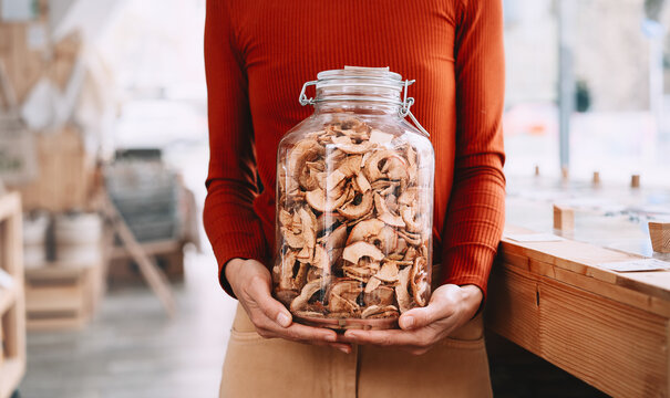 A happy caucasian female customer in a terracotta sweater holds a large glass jar of dried apples inside a bulk shop. Sustainable consumer choices and low waste lifestyle concept.