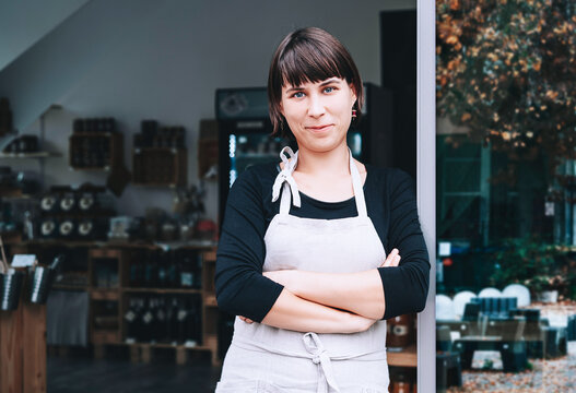 A confident caucasian female small business owner stands with crossed arms at the entrance of her local grocery shop. Authentic sustainable retail and successful entrepreneurship concept.
