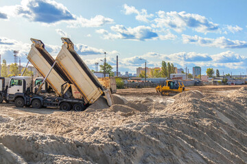 Two heavy tipper dump trucks unloading sand on a construction site with a bulldozer leveling ground in the background. © DmitryRider