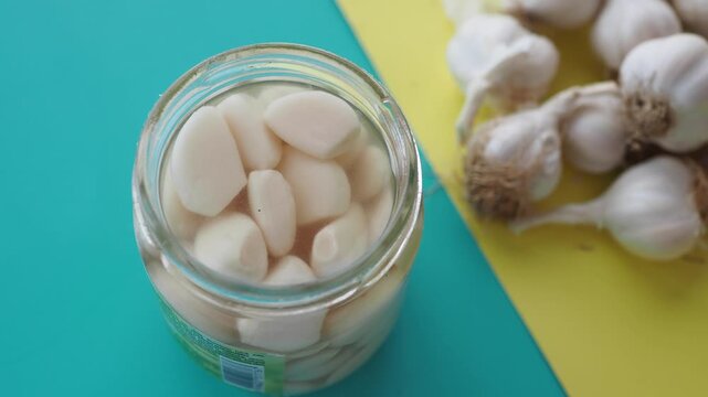 Pickled garlic cloves in glass jar and fresh whole garlic bulbs on yellow and blue background