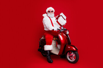 Naklejka premium Santa Claus in sunglasses rides a red scooter holding a wrapped gift against a red background
