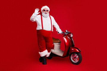 Naklejka premium Santa Claus waving while standing next to a red scooter on a vibrant red background, playful Christmas portrait with sunglasses and festive outfit