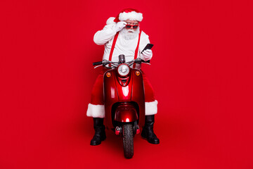 Naklejka premium Santa Claus on red scooter wearing sunglasses and checking smartphone for modern holiday deliveries and playful Christmas spirit