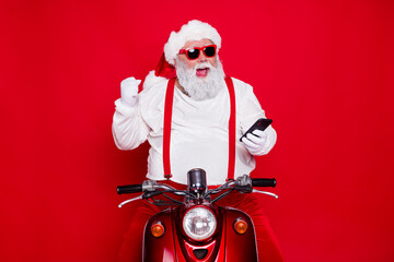 Naklejka premium Santa Claus on a red scooter wearing sunglasses and suspenders celebrates Christmas while checking a smartphone, cheerful holiday portrait with modern twist and playful festive energy