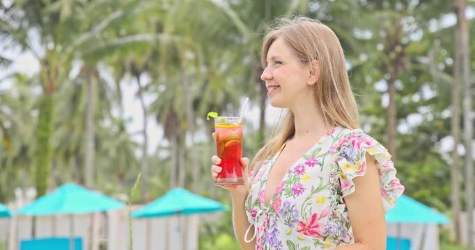 Happy young woman holding cold tropical cocktail at luxury swimming pool resort with palm trees. Smiling female tourist enjoying refreshing beverage on summer vacation.