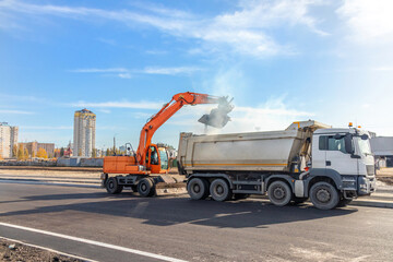 Orange wheeled excavator loading earth material into a heavy white dump truck on a new asphalt road during infrastructure construction. © DmitryRider