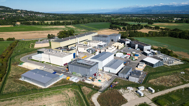 Aerial view of the European Center for Nuclear Research CERN facility with industrial buildings and surrounding green fields in Cessy, Auvergne-Rhone-Alpes, France.