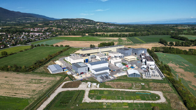 Aerial view of the European Center for Nuclear Research CERN facility buildings surrounded by green fields and agricultural land in Cessy, Auvergne-Rhone-Alpes, France.