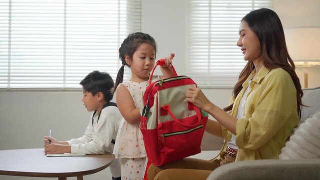 Asian mother helping daughter with her backpack before school while son does homework in the living room
