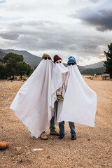 Fototapeta premium Three children dressed as ghosts for Halloween, wearing white sheets and hats. They stand together in a rural area with mountains in the background.