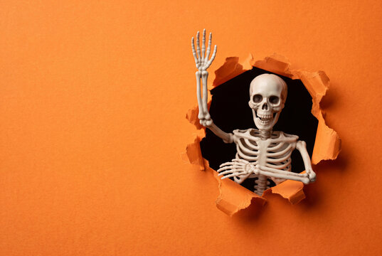 Skeleton waving from a hole in orange paper background