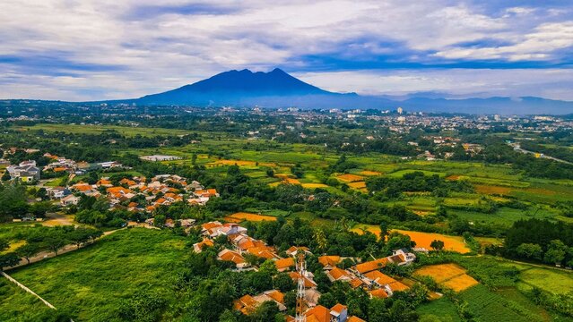 Aerial view of the Sentul area featuring lush green rice fields, residential houses with orange roofs, and a mountain backdrop in Babakan Madang, West Java, Indonesia.