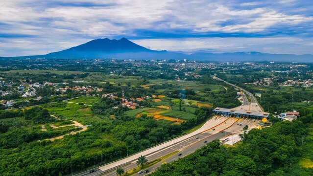 Aerial view of the Sentul toll gate surrounded by lush green vegetation and residential areas with a mountain in the background in Babakan Madang, West Java, Indonesia.