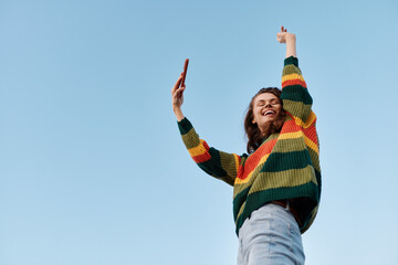 Joyful woman stands outdoors, wearing a vibrant striped sweater, arms raised in celebration, against a clear blue sky. A candid moment of happiness and energetic mood. © SHOTPRIME STUDIO