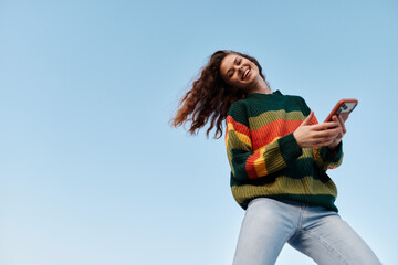 Happy young woman with curly hair in striped sweater smiling and using smartphone against blue sky....