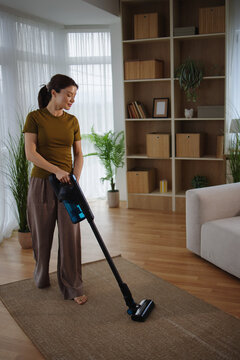 Woman cleaning living room carpet with a modern cordless stick vacuum cleaner, performing domestic chores and daily household tasks, maintaining a clean home environment