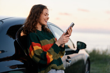 Obraz premium Smiling young woman in a colorful striped sweater leans on a car and uses her smartphone at sunset by the sea. Travel and leisure concept.