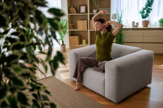 Woman stretching arms overhead and yawning while sitting comfortably on an armchair in a modern living room, embracing a tranquil morning routine at home