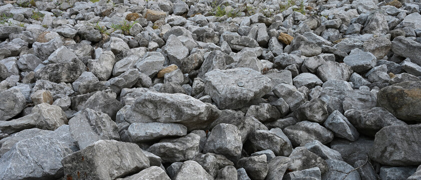 Riprap rock embankment with green grass in horizontal close-up, showing variegated gray stones of varied sizes and shapes, cracked rock surfaces, rough geological texture and uneven slope