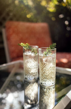 Two tall glasses of refreshing cocktail with ice and garnished with herbs placed on a glass table in a sunny outdoor setting with blurred background