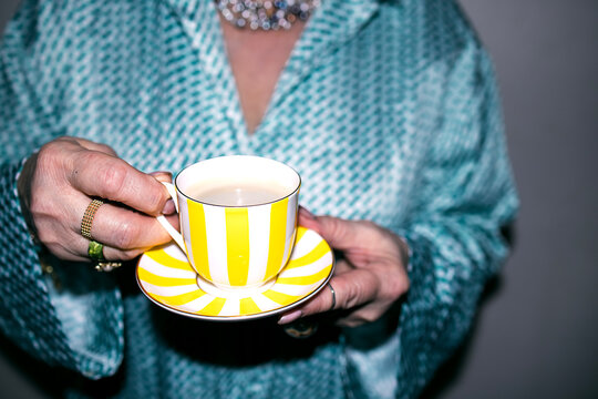 Woman holding a yellow and white striped teacup with saucer, wearing a turquoise patterned blouse and silver jewelry, against a neutral background